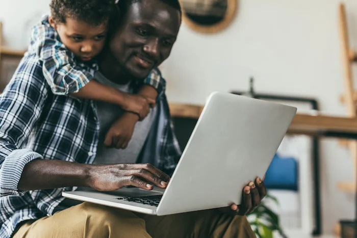 Father using laptop while young son hangs on shoulders